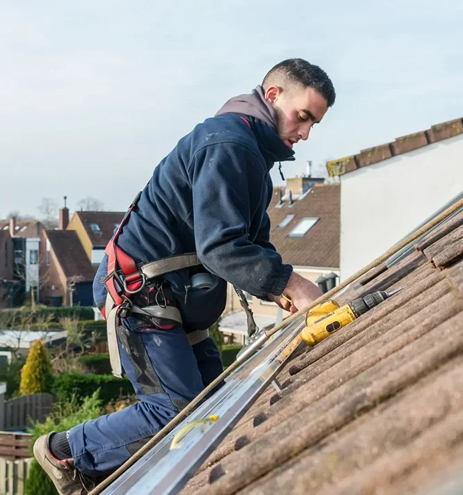 A worker in a safety harness adjusts roofing tiles on a sloped roof, with tools and a safety ladder nearby, against a suburban backdrop.