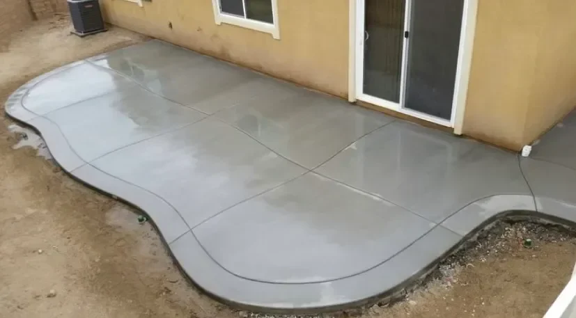 Freshly poured concrete patio with a smooth surface, featuring wavy edges, adjacent to a beige house with sliding glass doors.