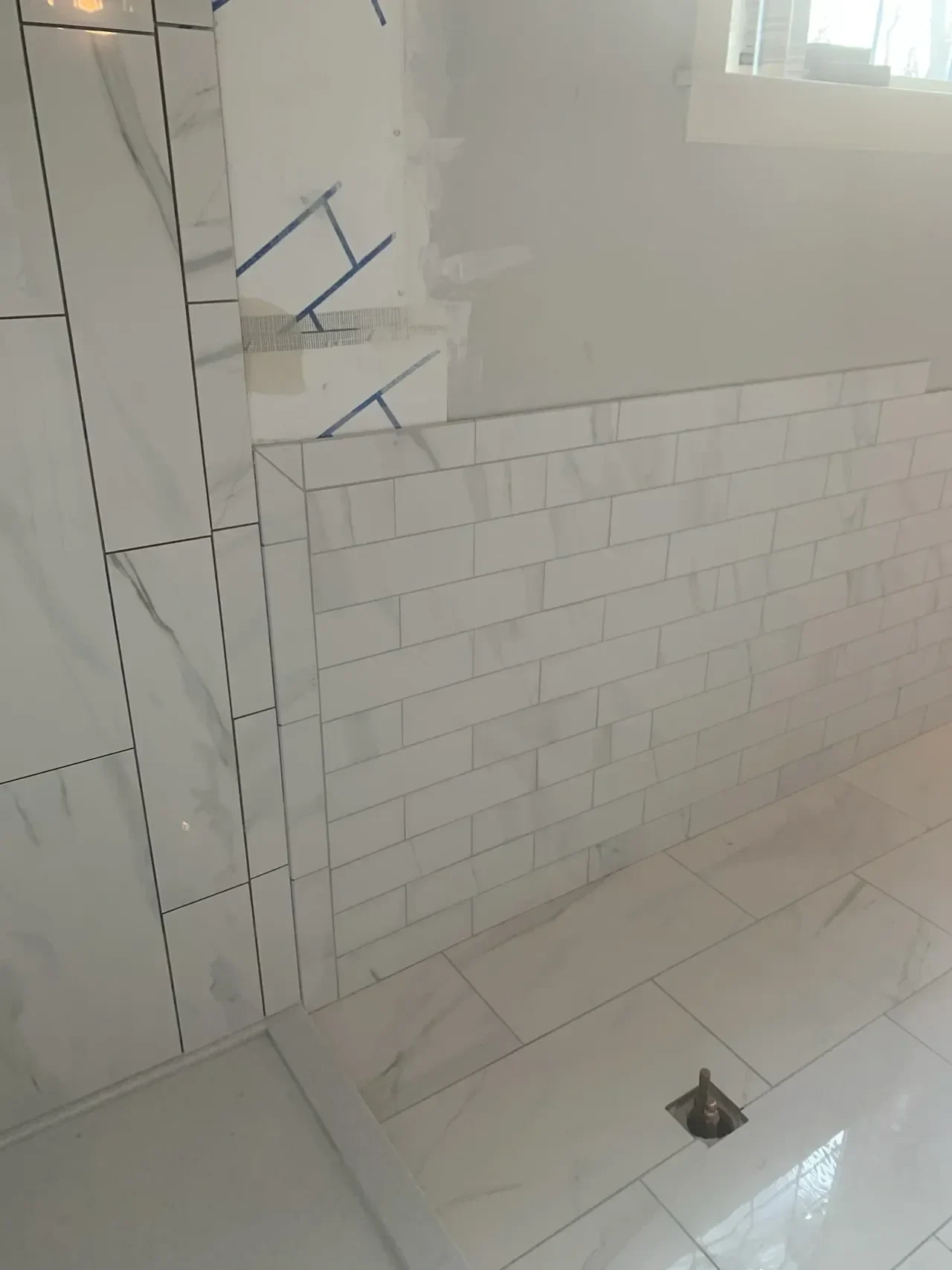 Interior view of a bathroom featuring white marble tiles in various sizes, with blue painter's tape on the wall, and a floor drain.