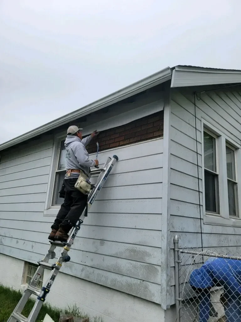 A man in a gray hoodie applies paint to the upper part of a house while balancing on a ladder next to a brick section.