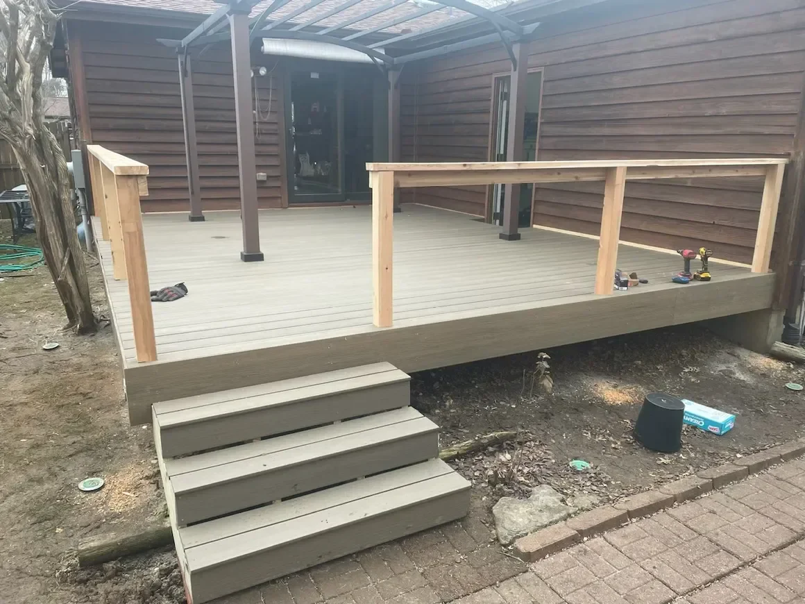 A newly constructed wooden deck with steps, surrounded by bare earth and tools, adjacent to a wooden house with a roofed pergola.