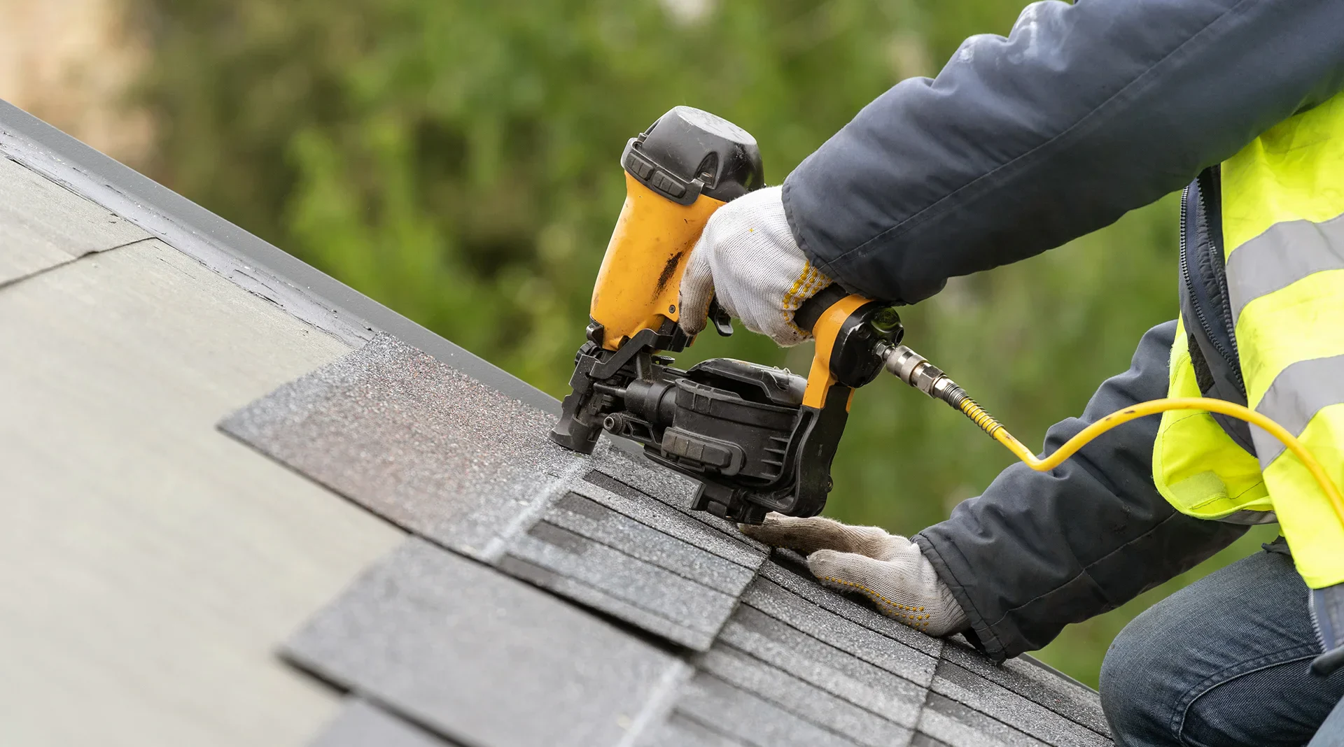 A worker in a yellow safety vest installs shingles on a roof using a pneumatic nail gun, surrounded by greenery.