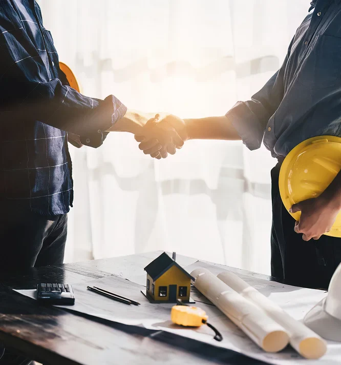 Two professionals shake hands over a construction table with blueprints, a calculator, and a small house model in bright, natural light.