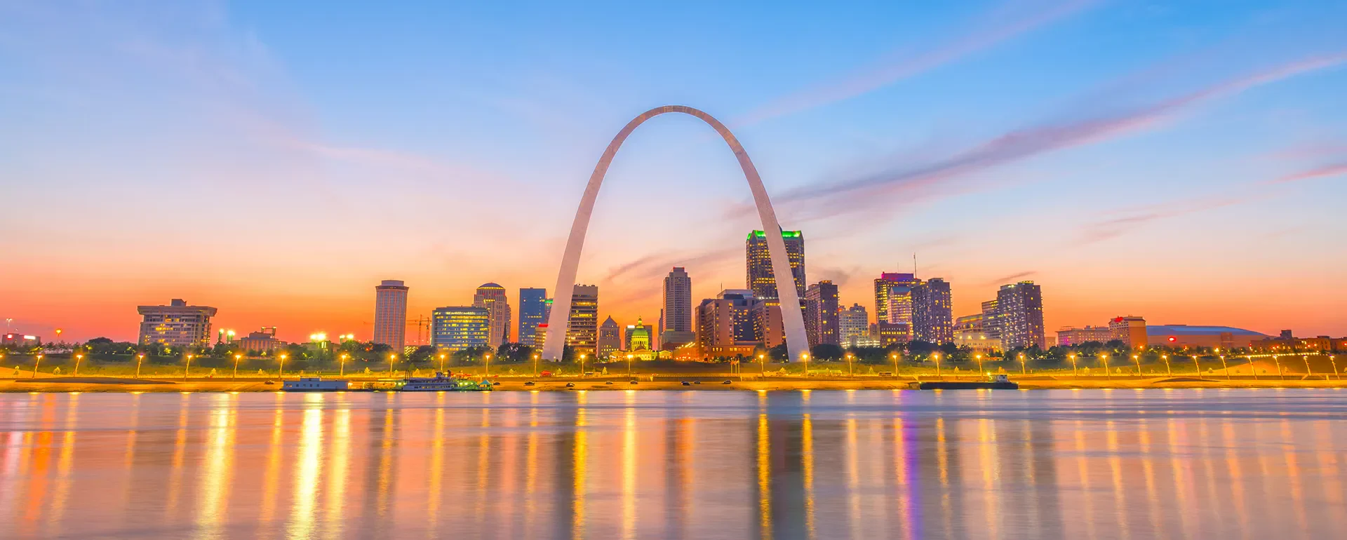 Sunset over St. Louis, featuring the iconic Gateway Arch and a shimmering river, with colorful city skyline lights reflecting on the water.