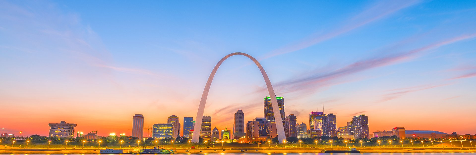 St. Louis skyline at sunset featuring the Gateway Arch, illuminated buildings, and colorful sky.